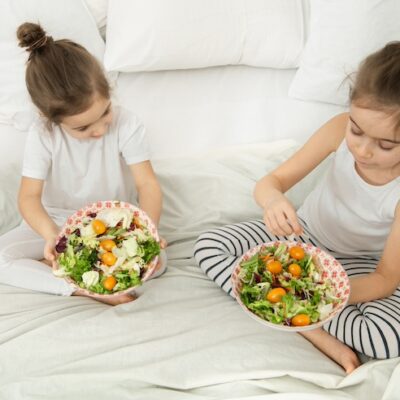Two girls sitting on a bed holding bowls of salad.