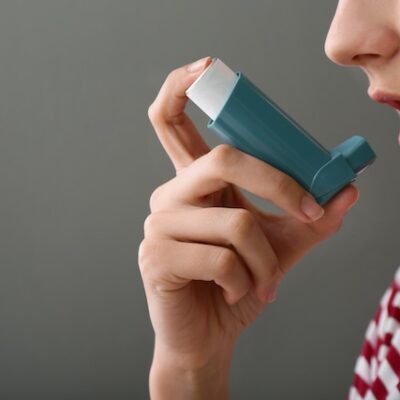 Young woman using inhaler against asthma on grey background.