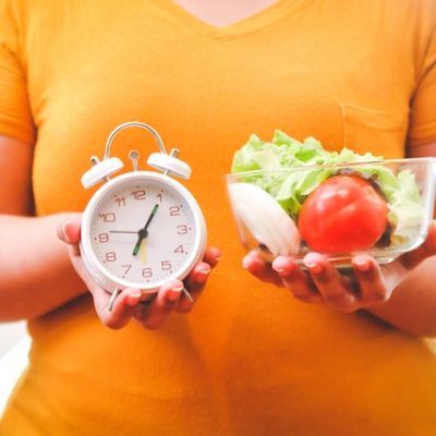 Woman in an orange t-shirt holding food in one hand and an alarm clock in the other.