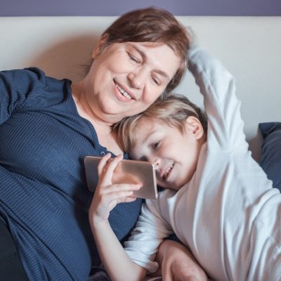Happy grandmother and her small grandson watching cartoons on smart phone in bedroom.