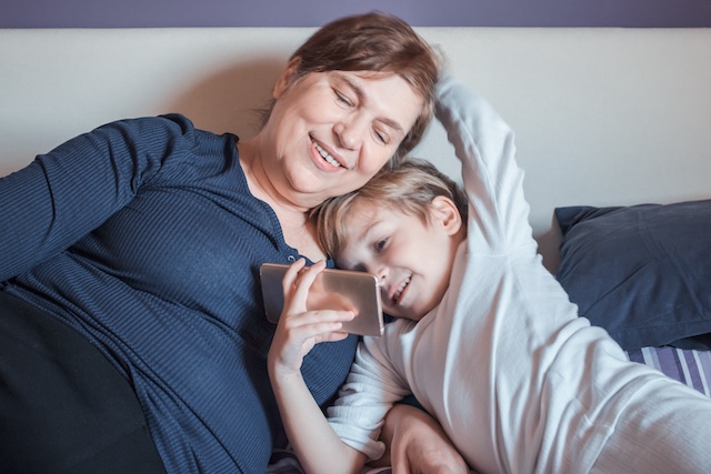 Happy grandmother and her small grandson watching cartoons on smart phone in bedroom.