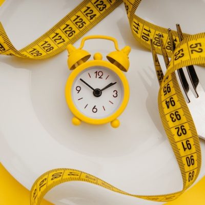 Yellow alarm clock on white plate with cutlery. Concept of intermittent fasting, lunchtime, diet and weight loss