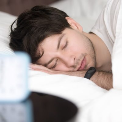 Young guy sleeping in bed next to an alarm clock.