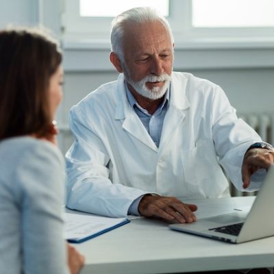 Mature doctor and his patient talking while reading medical record on a computer at doctor's office.