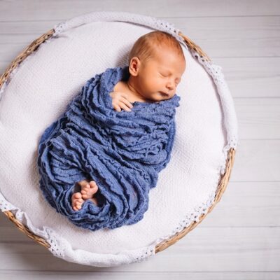 Newborn baby enveloped in blue scarf sleeps on white pillow.